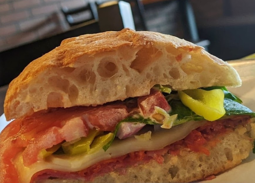 A close-up of a sandwich with layers of meat, cheese, tomatoes, spinach, and peppers on a crusty bread roll.